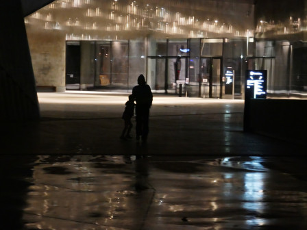 L'ombre d'une mère et de son enfant, sous une passerelle, la nuit, près de la Philarmonie de Paris. Au fond, un bâtiment illuminé.