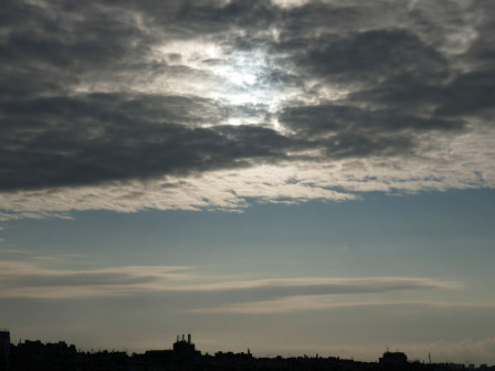 Le soleil aperçu dans une couche de nuages furieux, le ciel plus dégagé en dessous, les faites des toits parisiens, en bas.