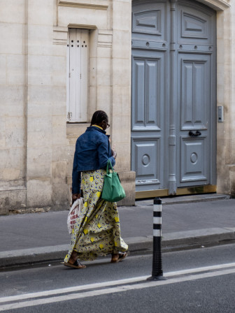 Une femme noire à tenue audacieuse, bleu jean, jupe vert pâle, sac vert vif, s'apprête à passer devant une porte bleu clair. on la voit de 3/4 dos.